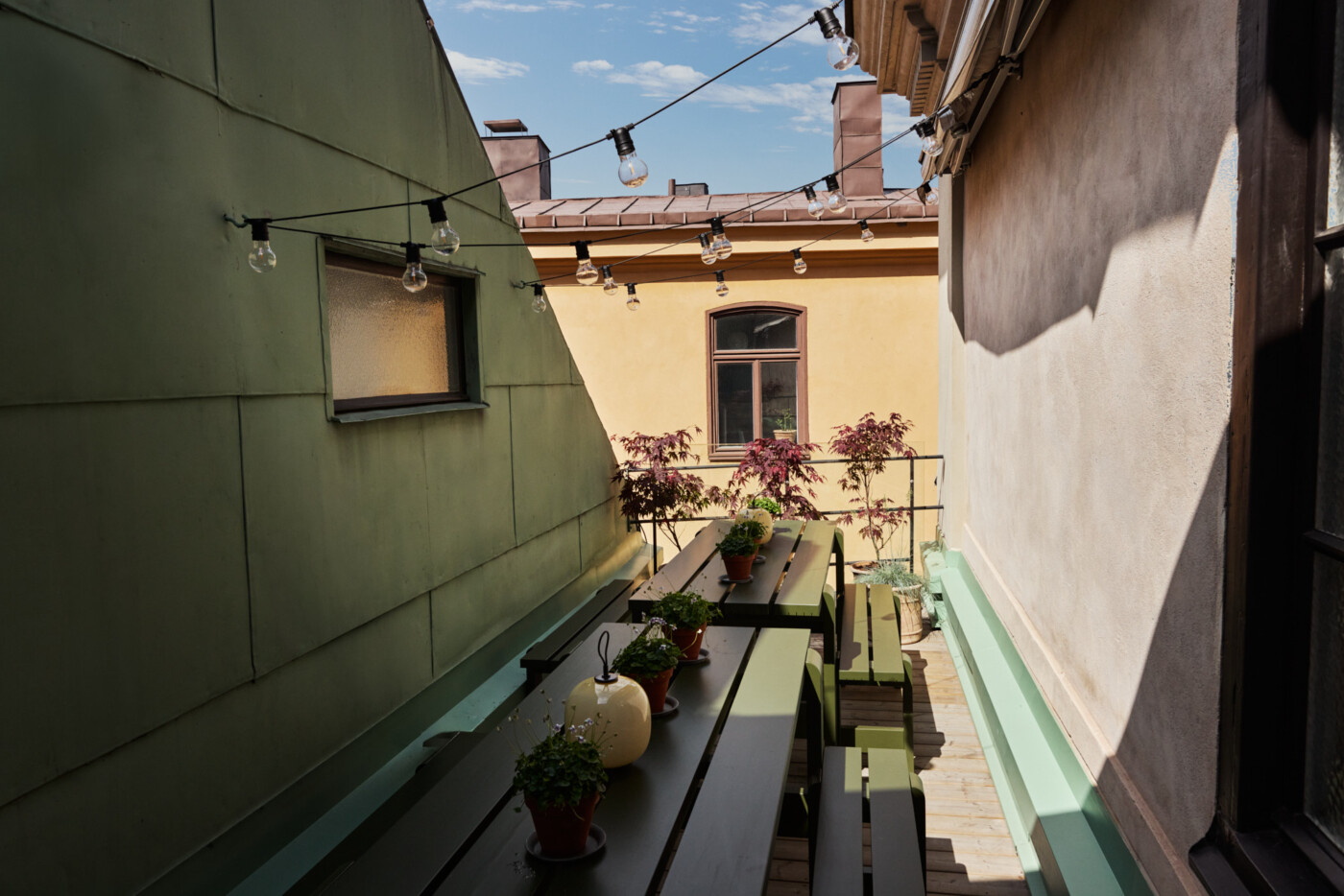 Balkongen på Okidoki Arkitekt's Stockholms kontor. Grönt bord och bänkar med blommor på bordet och ljuslingor mot en blå himmel